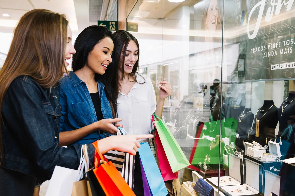 Três mulheres sorridentes fazendo compras em shopping center, olhando vitrine de joias com sacolas coloridas nas mãos.