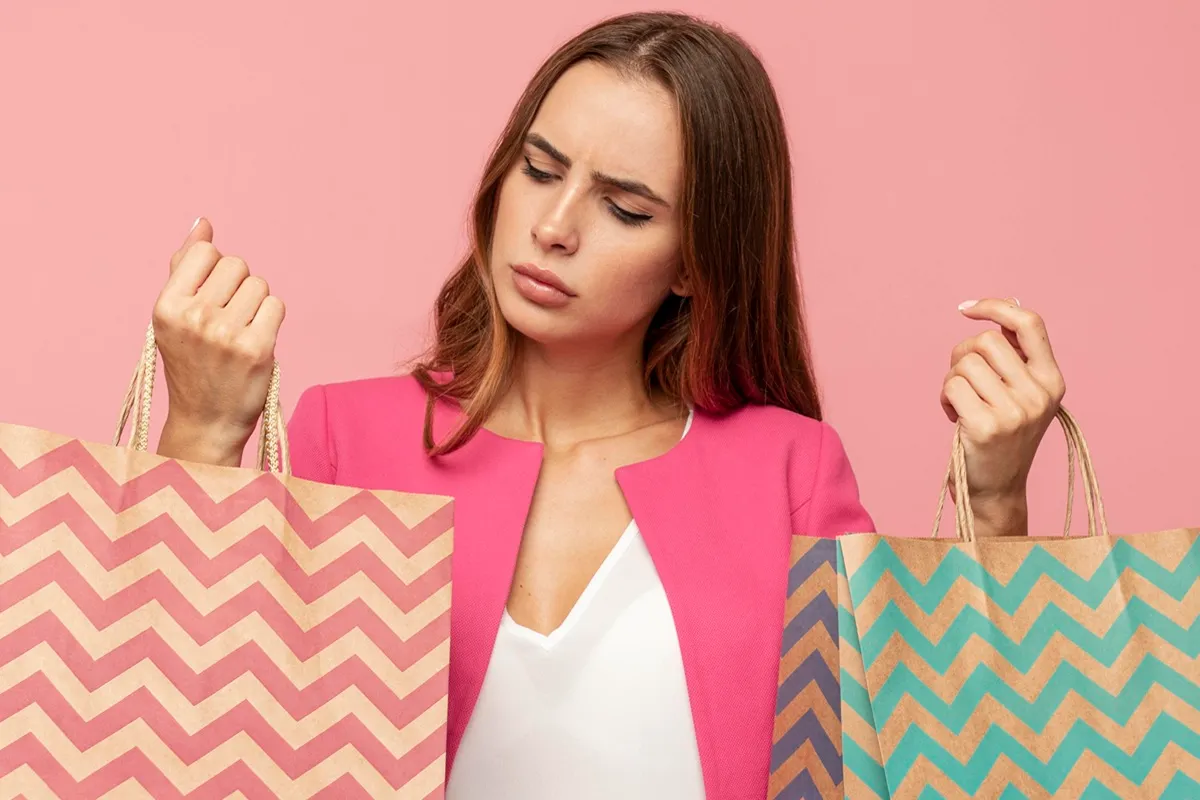 Mulher jovem de expressão confusa segurando duas sacolas de papel estampadas, uma em cada mão, vestindo blazer rosa e blusa branca, em frente a um fundo rosa sólido.