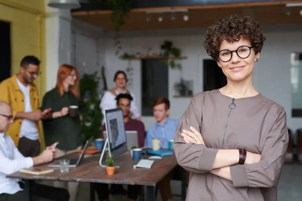 Mulher sorridente de braços cruzados com equipe trabalhando ao fundo em ambiente corporativo moderno.