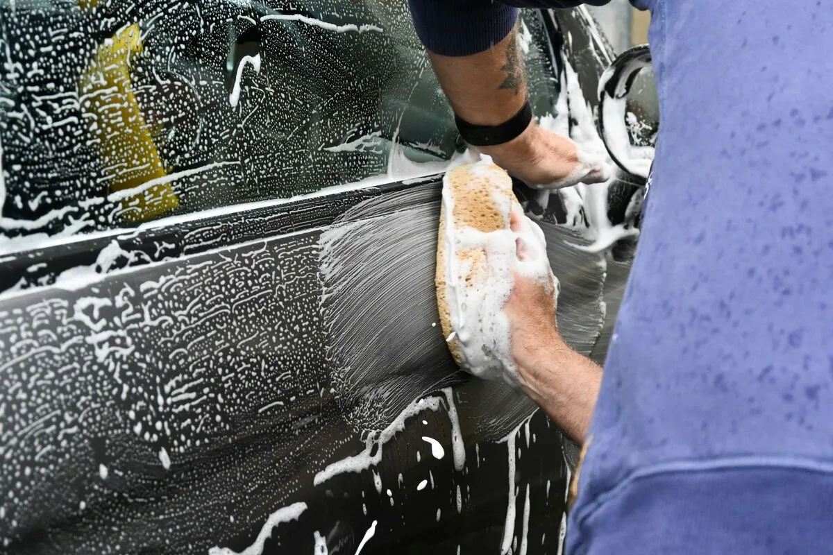 Pessoa lavando carro preto com esponja e sabão, detalhando a limpeza da lataria.