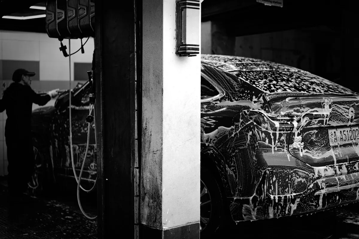 Carro sendo lavado com espuma em lava-jato coberto, imagem em preto e branco com funcionário ao fundo.