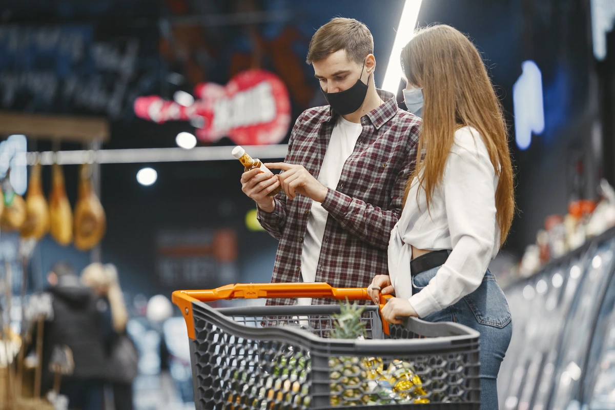 Casal usando máscaras enquanto faz compras em mercados atacadistas, analisando rótulo de produto com carrinho cheio.