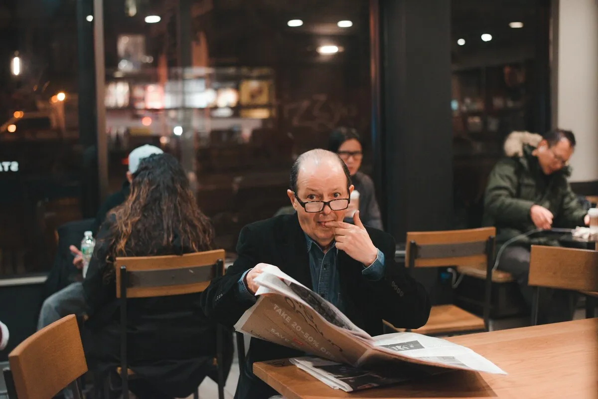 Homem de meia-idade lendo jornal em cafeteria moderna durante a noite, usando óculos e fazendo expressão curiosa, com outras pessoas ao fundo interagindo.