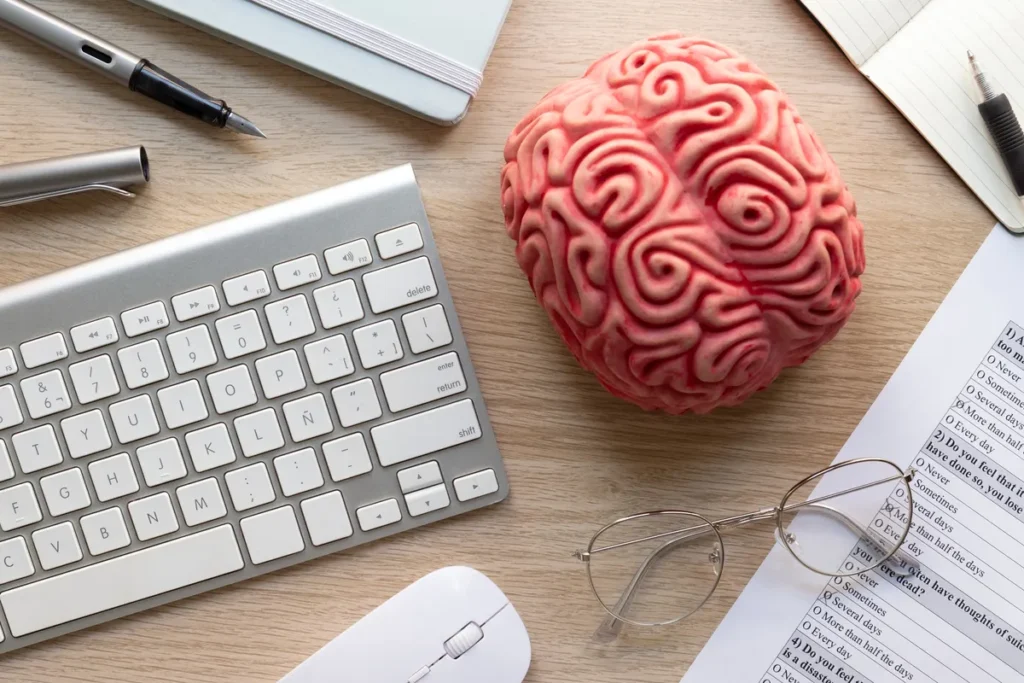 Cérebro de plástico sobre mesa de escritório ao lado de teclado, mouse, óculos e caderno, simbolizando neuromarketing, criatividade e produtividade no trabalho digital.