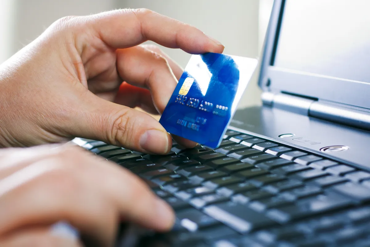 Pessoa segurando cartão de crédito azul enquanto digita em notebook para realizar compra online