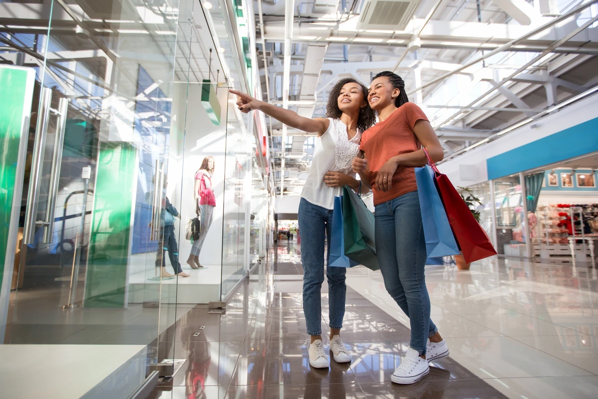 Duas mulheres felizes com sacolas de compras andando em shopping moderno e observando vitrines de lojas.
