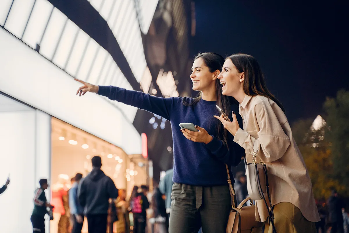 Duas mulheres sorrindo e apontando para vitrine iluminada de loja durante passeio noturno em área comercial movimentada.