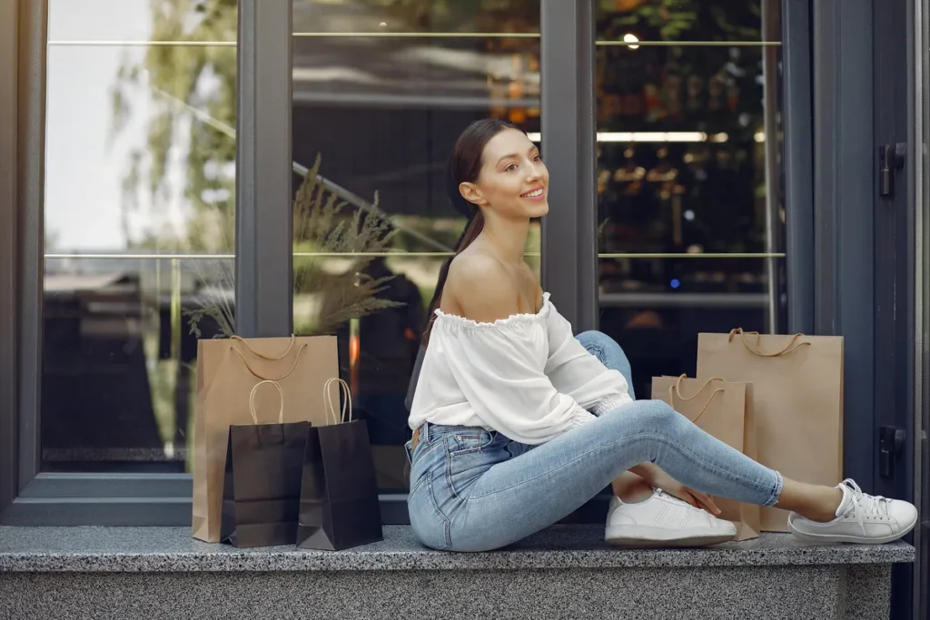 Mulher jovem, sorridente, sentada do lado de fora de uma loja com várias sacolas de compras ao lado, possivelmente em um outlet.