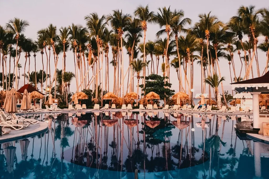 Imagem de uma piscina de resort ao entardecer, com palmeiras altas e guarda-sóis de palha ao fundo. As palmeiras e estruturas de palha refletem na água calma da piscina, criando uma atmosfera relaxante.