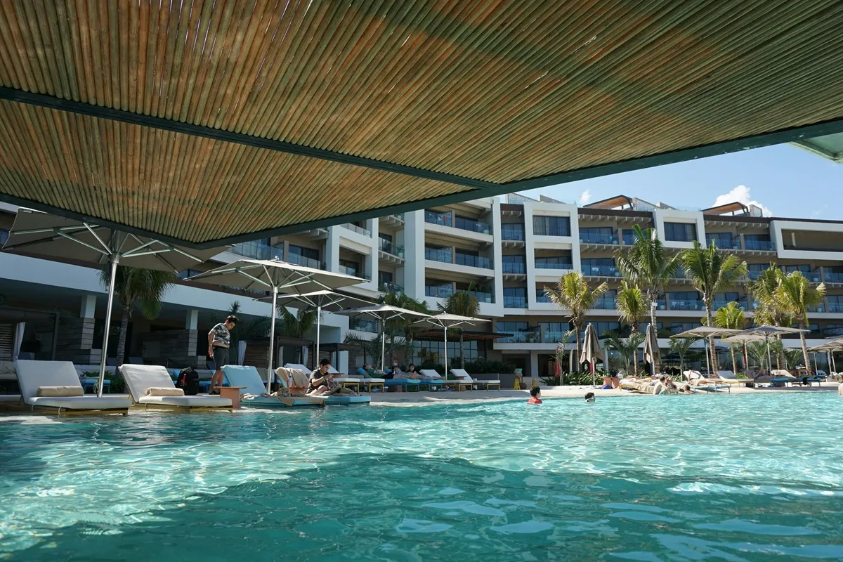 Imagem de uma piscina de resort com espreguiçadeiras sob guarda-sóis, vista para um hotel moderno e um céu claro. A piscina exibe águas calmas, com alguns hóspedes relaxando e nadando.