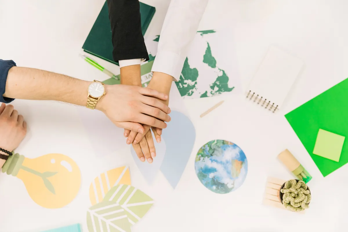Mãos sobrepostas de pessoas em sinal de união e colaboração sobre mesa com materiais de sustentabilidade, mapas e ícones ecológicos, representando responsabilidade social e ambiental.