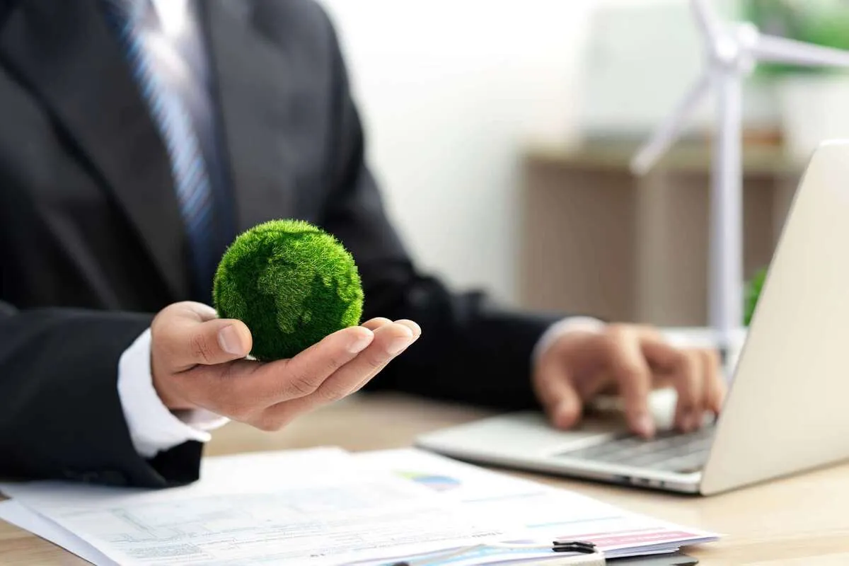 Empresário segurando um globo verde de grama que representa sustentabilidade, enquanto trabalha no notebook, com turbina eólica ao fundo e documentos na mesa.