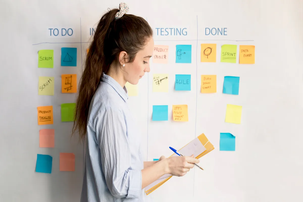 Mulher analisando tarefas em quadro Kanban com post-its organizados por etapas de um projeto ágil, incluindo Scrum, Agile e Sprint