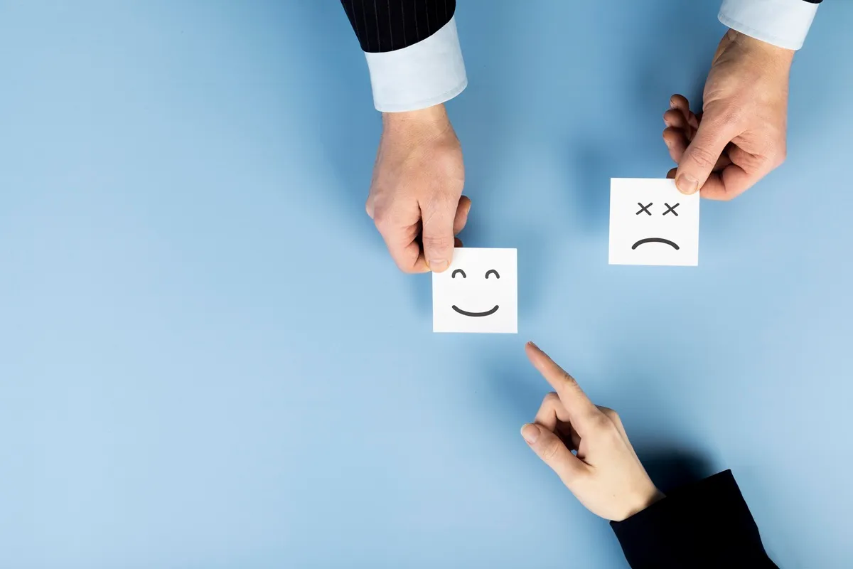 Mãos segurando cartões com carinhas feliz e triste, representando feedback positivo e negativo, sobre fundo azul claro.