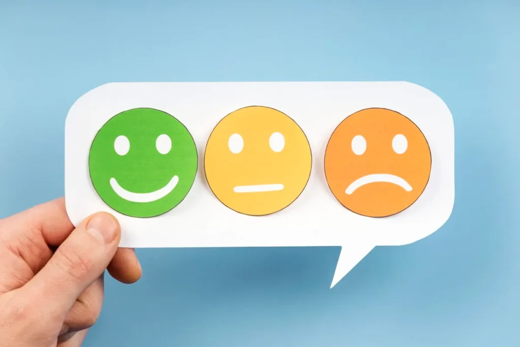 Três emojis coloridos representando níveis de satisfação: rosto verde sorridente, rosto amarelo neutro e rosto laranja triste, dentro de balão de fala segurado por uma mão, sobre fundo azul.