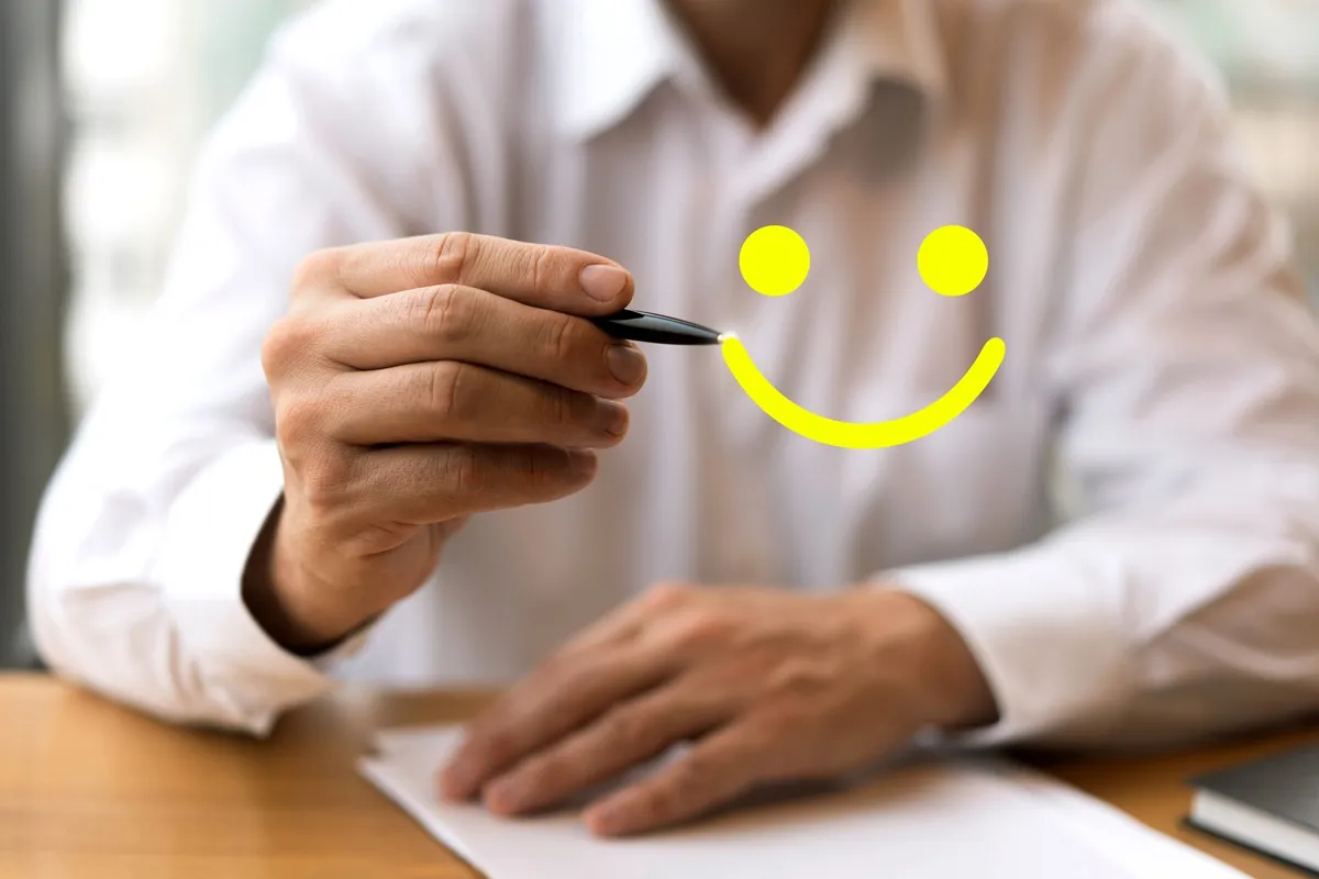 Homem com camisa branca segurando caneta e desenhando um smiley amarelo em frente a uma folha de papel, representando feedback positivo.