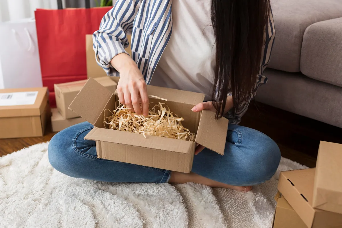 Mulher abrindo caixa de papelão no chão cercada por outras embalagens, representando momento de unboxing e experiência pós-compra no e-commerce.