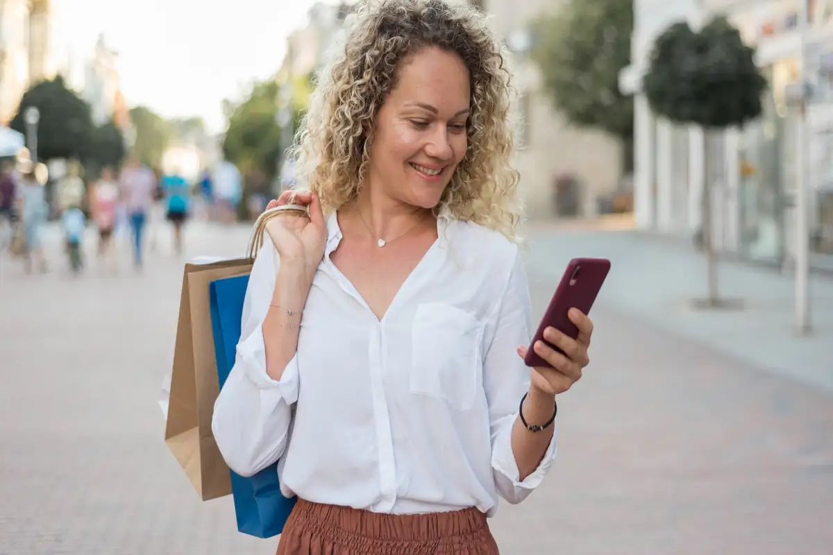 Mulher sorridente olhando o celular enquanto carrega sacolas de compras, representando experiência de compra omnichannel e usabilidade mobile.