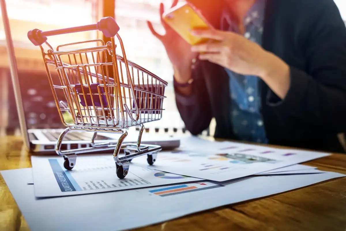 Carrinho de compras em miniatura sobre gráficos de desempenho, com pessoa usando smartphone ao fundo, representando análise de dados e usabilidade no e-commerce.