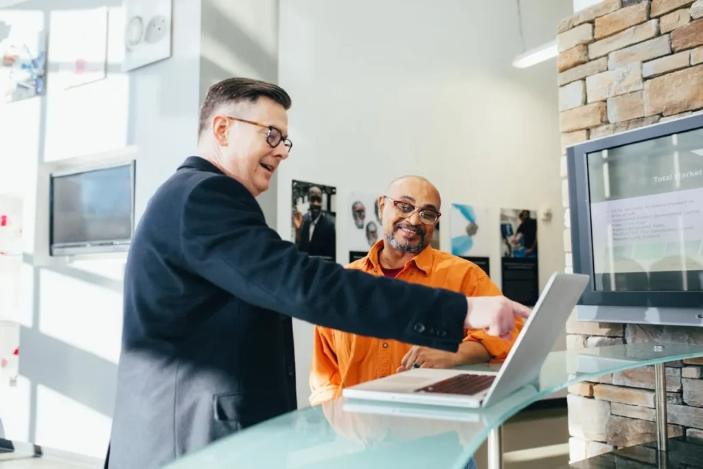 Vendedor proativo apontando para um laptop, apresentando soluções a um cliente em ambiente de negócios moderno.