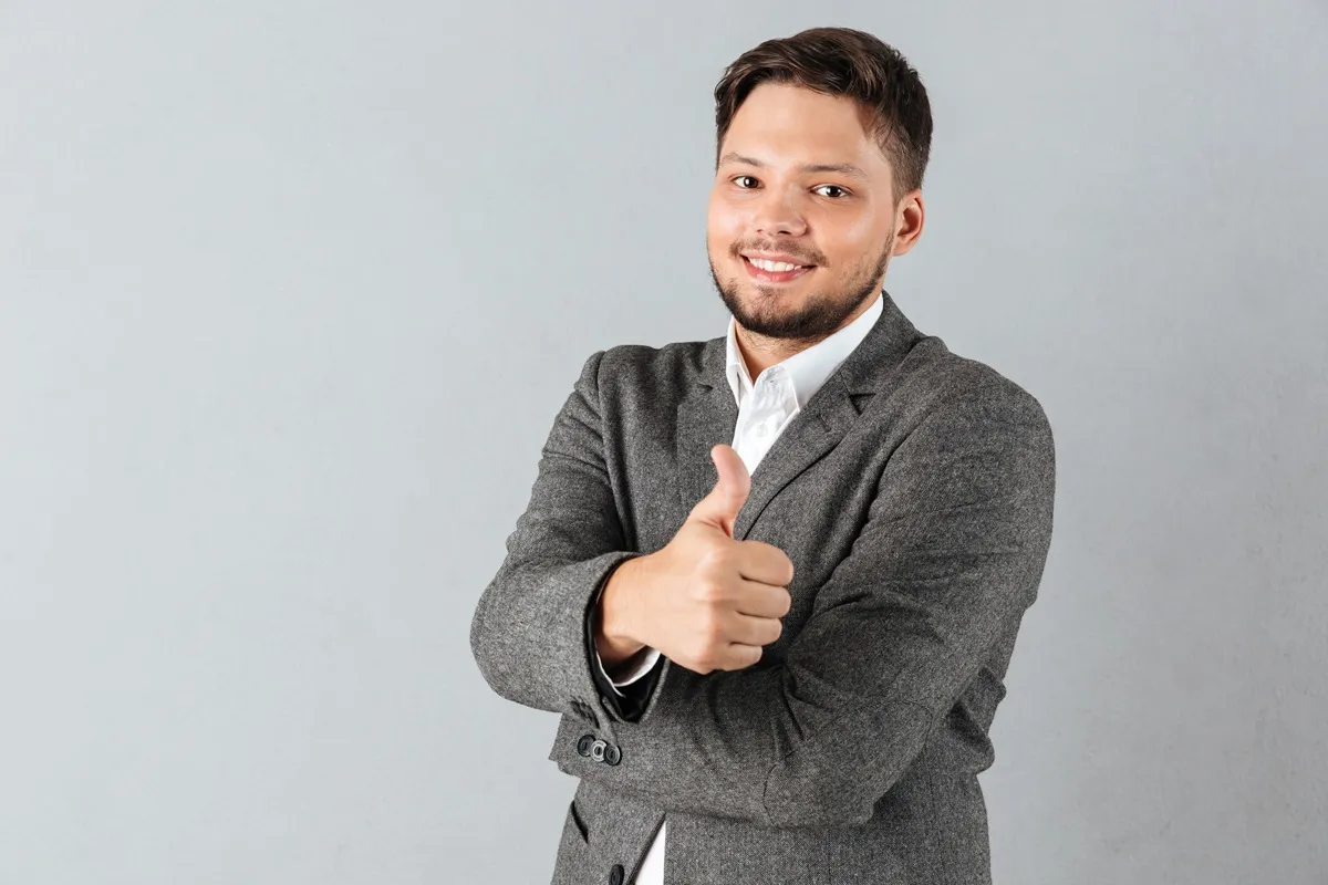 Homem jovem com barba curta e cabelo castanho, usando blazer cinza e camisa branca, sorrindo e fazendo sinal de "joinha" com o polegar levantado em frente a um fundo cinza claro.