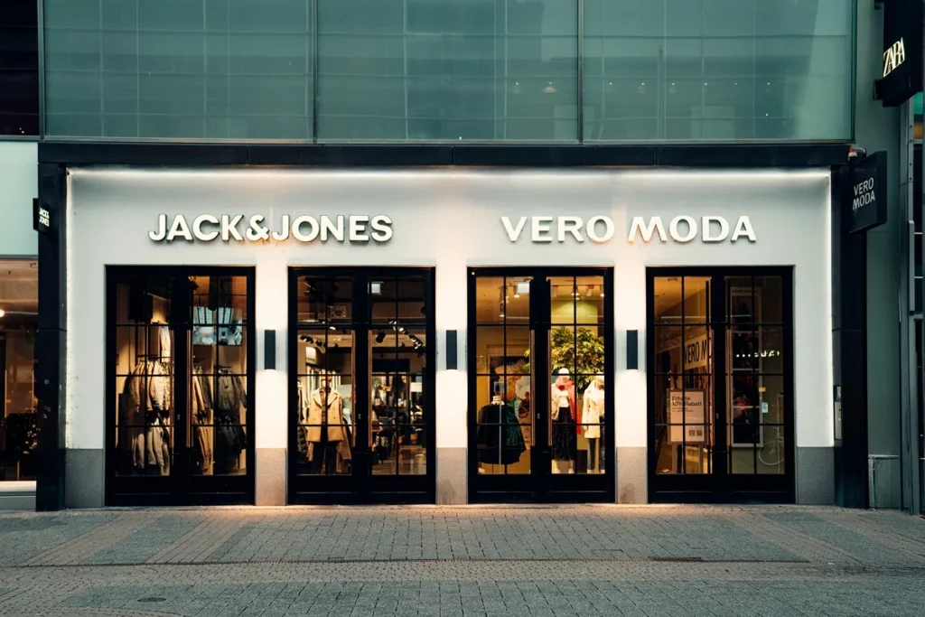 Fachada de duas lojas de roupas, Jack & Jones e Vero Moda, com portas de vidro, vitrines iluminadas exibindo manequins e decoração clean