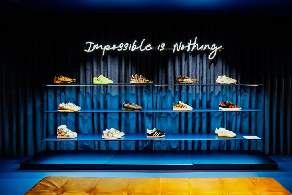 Vitrine interna com três prateleiras azuis exibindo diversos modelos de tênis e um letreiro neon branco escrito ‘Impossible is Nothing’”