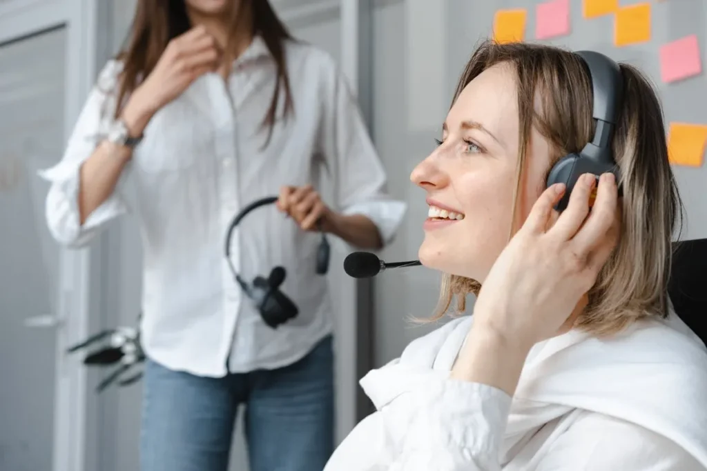 Mulher sorridente com fones de ouvido e microfone atendendo a uma chamada em um centro de atendimento ou escritório, com uma colega ao fundo, representando atendimento ao cliente e Customer Experience (CX).
