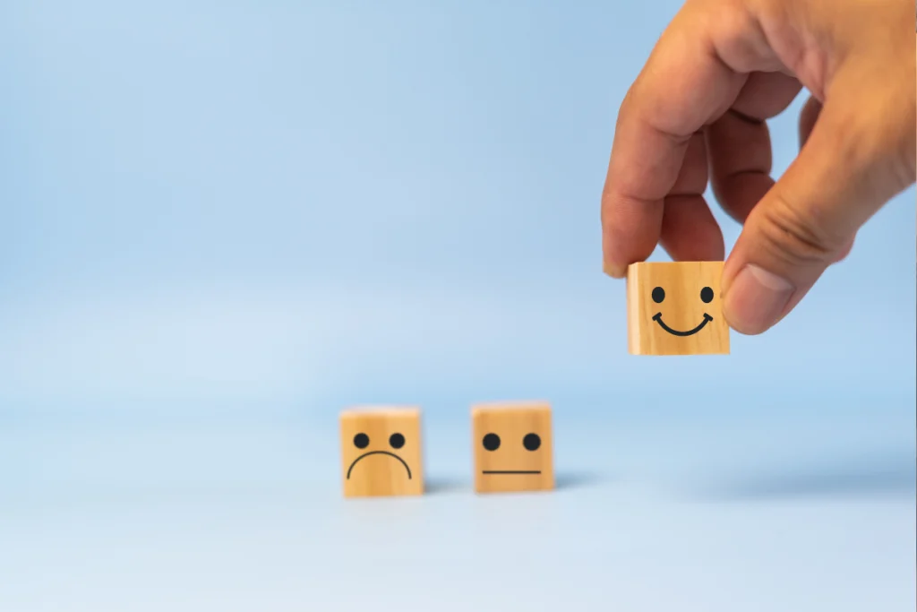 Mão segurando bloco de madeira com rosto sorridente ao lado de blocos com expressões triste e neutra, simbolizando níveis de satisfação.
