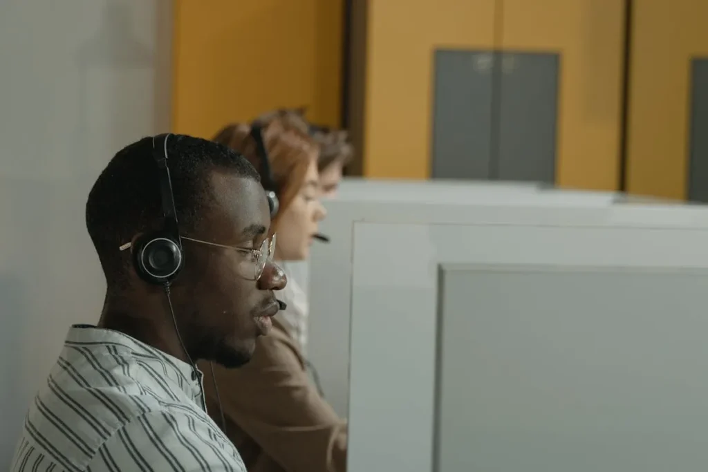 Homem negro usando óculos e fone de ouvido com microfone, sentado em uma baia de um call center, concentrado em seu trabalho.