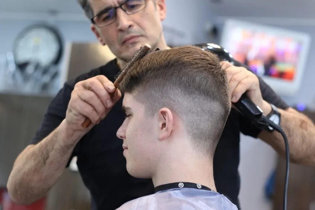 Barbeiro profissional cortando o cabelo de um jovem, utilizando secador e escova, em um ambiente de salão de beleza. O foco está na qualidade do serviço, visando a satisfação do cliente e um NPS promotor.