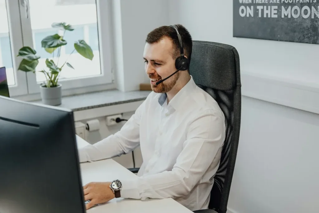 Um homem de barba e fone de ouvido está sorrindo enquanto trabalha em um computador. Ele parece estar oferecendo suporte ao cliente para a TMA, demonstrando um atendimento online dedicado e profissional.