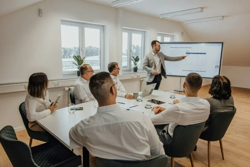 Um grupo de profissionais de negócios está em uma sala de reunião moderna. Um homem em pé, no centro, aponta para uma tela grande que exibe gráficos e dados, provavelmente resultados ou funcionalidades da TMA. Ele está apresentando para outros cinco colegas sentados à mesa, que estão atentos à demonstração da TMA.