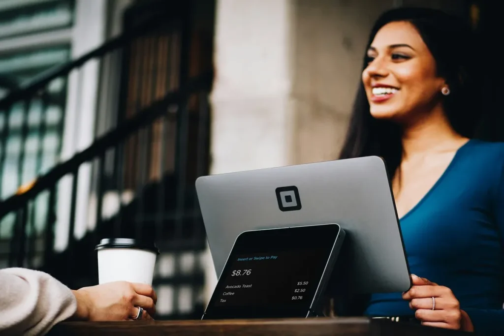 Uma barista sorridente opera um sistema de ponto de venda da TMA em um café, enquanto um cliente segura um copo de café descartável. A tela do sistema mostra um pedido de "Avocado Toast", "Coffee" e "Tax", totalizando $8.76, demonstrando a eficiência da TMA em estabelecimentos de alimentos e bebidas.