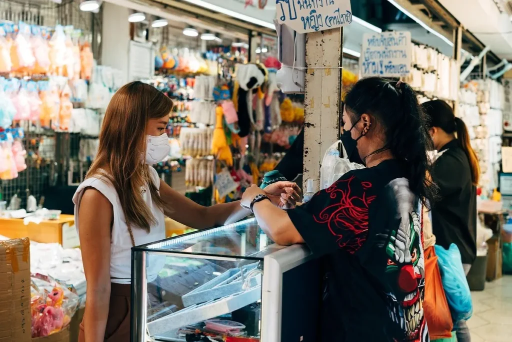  Duas mulheres usando máscaras trocando um produto em um balcão de loja, com prateleiras de mercadorias ao fundo, ilustrando a interação e a experiência atitudinal no varejo.