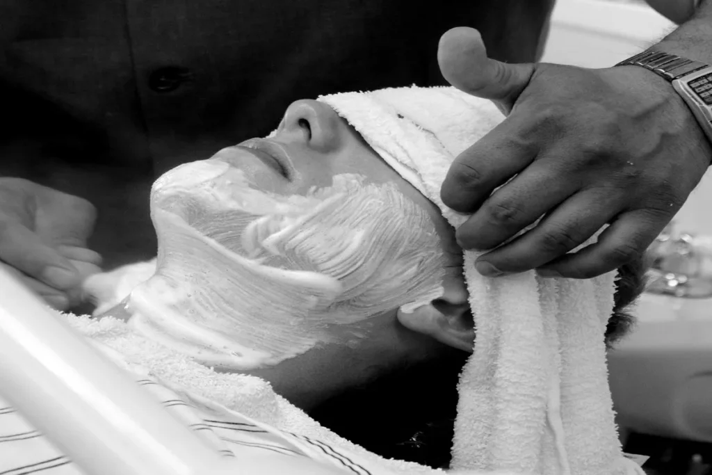 Um homem recebendo uma toalha quente e espuma de barbear no rosto em uma barbearia, representando um serviço atencioso e a experiência do cliente.