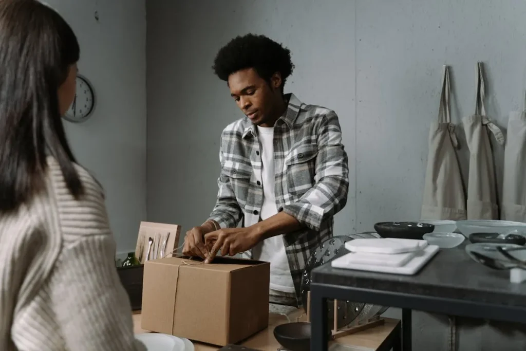 Homem embalando caixa para envio, com mulher observando, representando transações de e-commerce B2B e B2C.