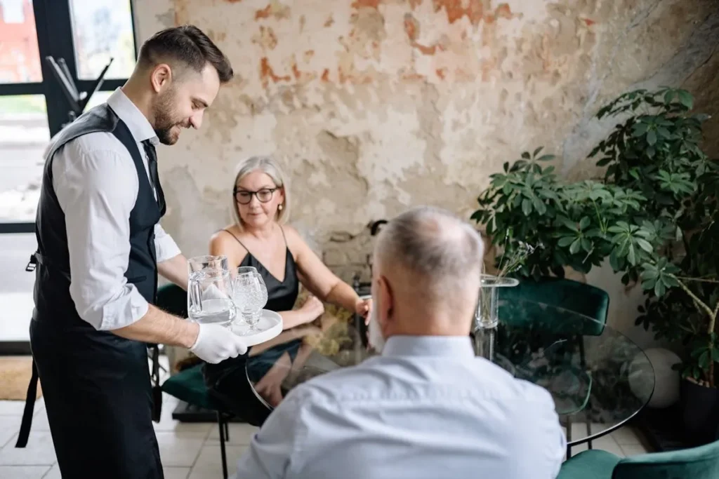 Garçom servindo água para um casal em restaurante rústico, ilustrando a interação em modelos de negócios B2C e B2B.