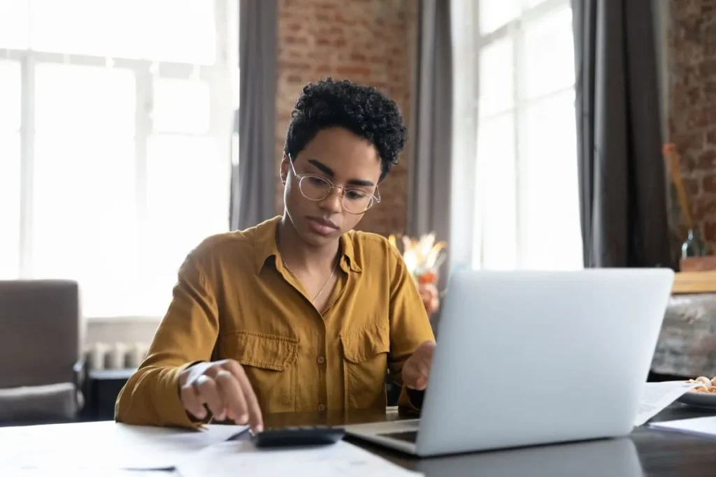 Jovem séria, de óculos, usando um laptop e uma calculadora em uma mesa de escritório rústica, analisando dados financeiros para benchmarks ou planejamento.