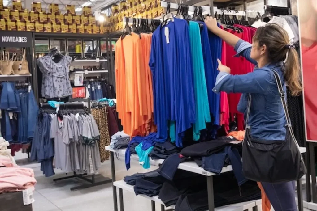 Mulher navegando em um expositor de roupas de moda em uma loja de comércio varejista, segurando uma peça de roupa azul. A loja tem várias prateleiras com roupas em exposição, calçados e um letreiro de "Novidades".