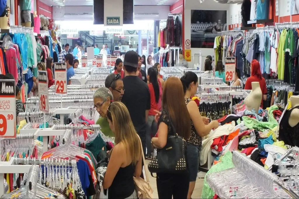 Clientes em uma loja de departamento movimentada, navegando por araras de roupas, representando o comércio varejista.
