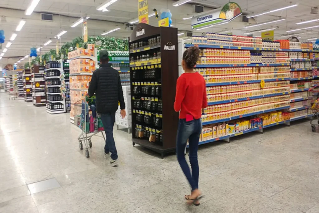 Casal andando em corredor de supermercado, com carrinho de compras, em um ambiente de comércio varejista. Prateleiras cheias de produtos, com foco na seção de temperos e alimentos.