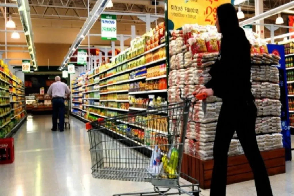 Uma cliente empurra um carrinho de compras em um supermercado movimentado, representando o comércio varejista de alimentos.