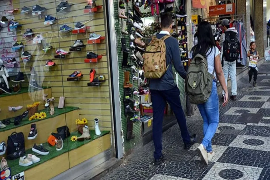 Clientes com mochilas caminham na calçada de uma rua, passando por lojas de comércio varejista de calçados.