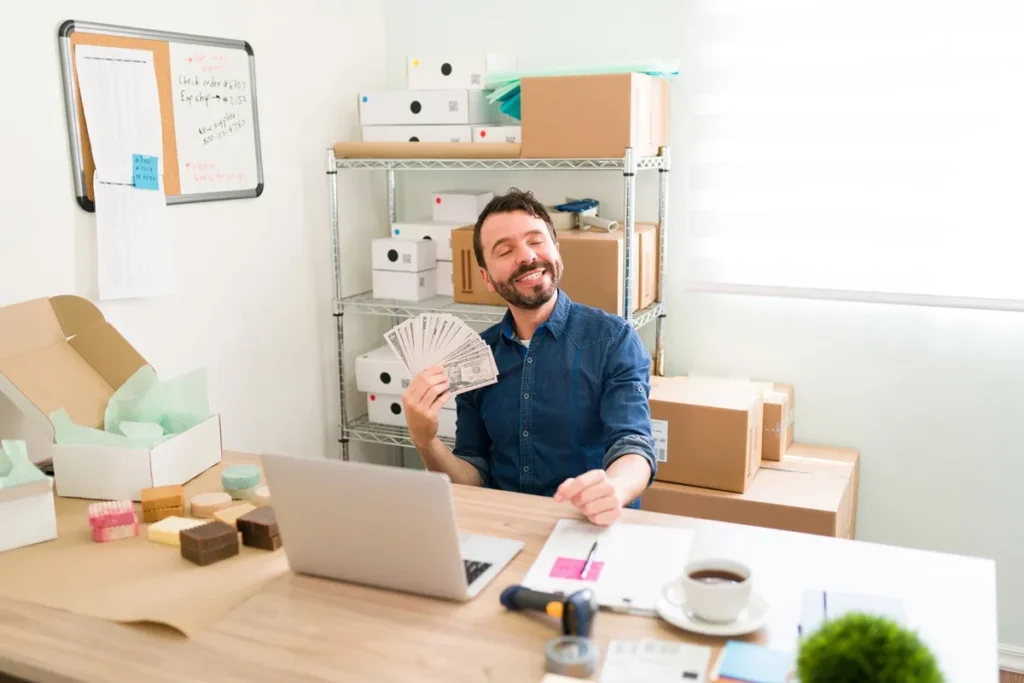 Homem sorridente segurando notas de dinheiro em um escritório com caixas e produtos embalados, simbolizando sucesso em vendas online.