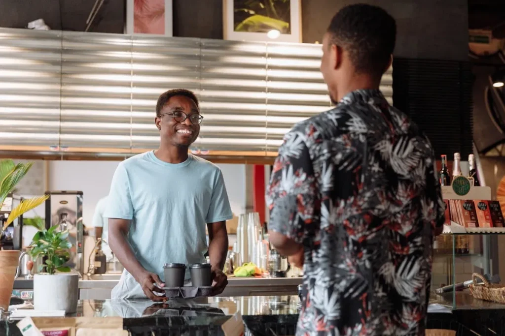 Homem sorridente, com óculos e camiseta azul clara, entregando duas bebidas de café em um suporte para um cliente em um balcão de cafeteria. A cena transmite credibilidade e bom atendimento