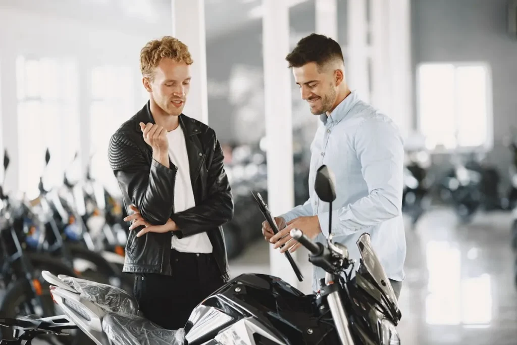 Dois homens em uma concessionária de motocicletas, um deles vestindo jaqueta de couro preta e o outro camisa azul clara, examinando uma motocicleta preta. O homem de camisa azul segura uma prancheta e parece estar explicando detalhes da moto para o outro homem.