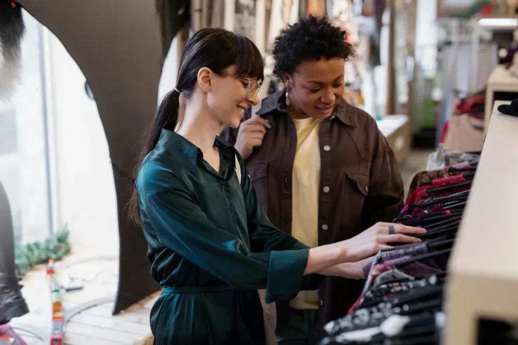 Duas mulheres em uma loja de roupas, uma delas, sorrindo e usando óculos, está folheando cabides cheios de vestuário, enquanto a outra observa, representando a experiência de compra em "departamentos" de moda.