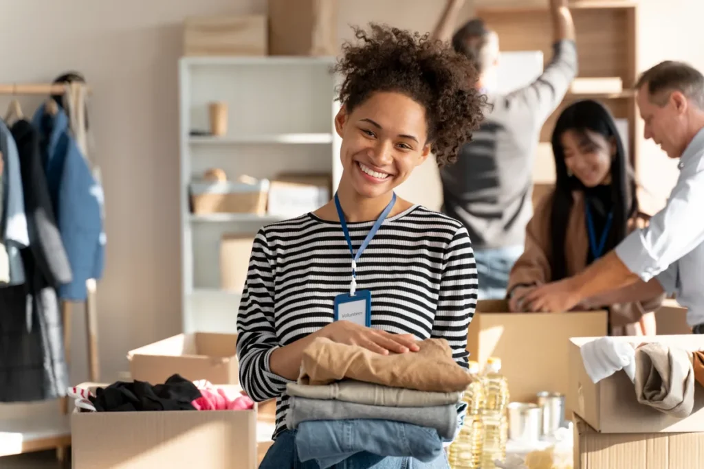 Uma jovem voluntária sorridente segurando uma pilha de roupas dobradas em um centro de doações, com outras pessoas trabalhando em segundo plano classificando itens, representando a organização de "departamentos" em um trabalho voluntário ou centro de distribuição.