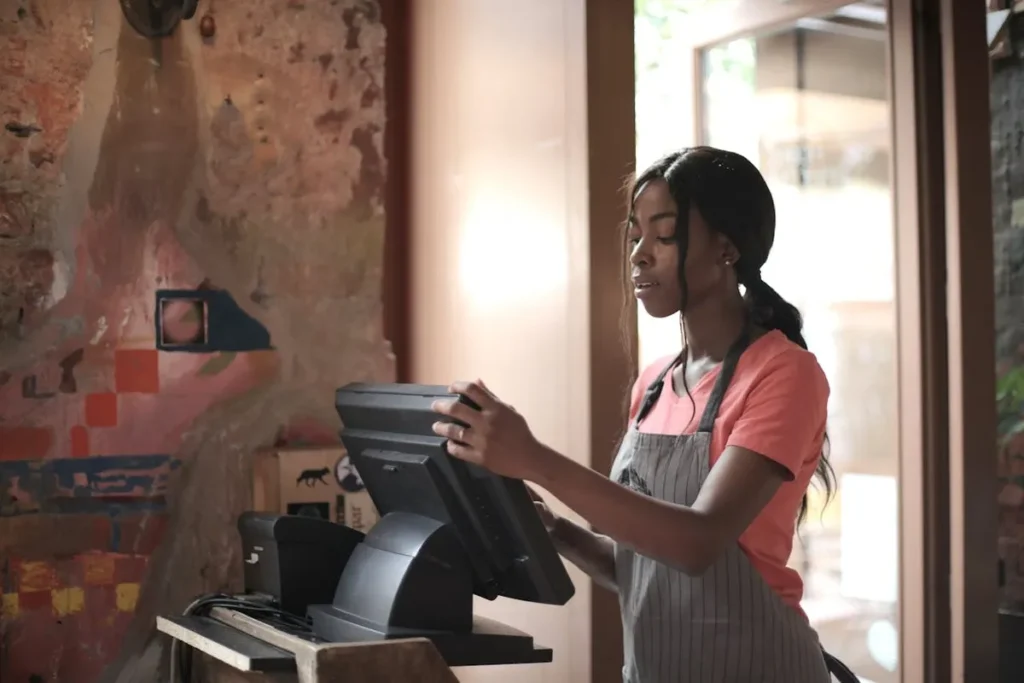 Uma jovem atendente negra, vestindo avental e camiseta, utiliza um sistema de ponto de venda (PDV) em um estabelecimento comercial, evidenciando a eficiência operacional na gestão de pedidos e pagamentos.